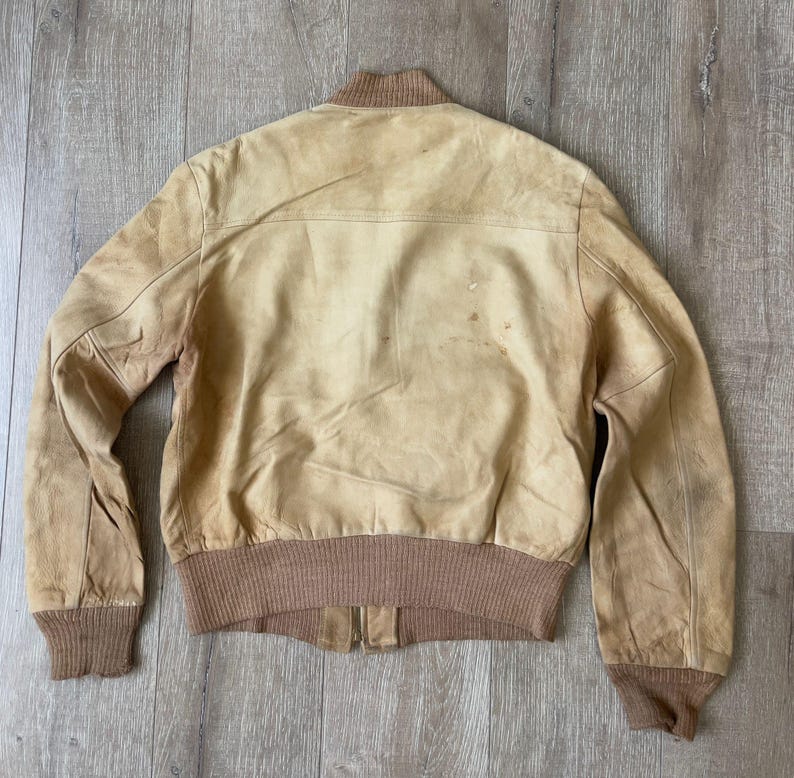 May include: A tan leather bomber jacket with a brown knit collar, cuffs, and waistband. The jacket has a zipper closure and visible wear and tear, giving it a vintage look. The jacket is laid flat on a wooden surface.