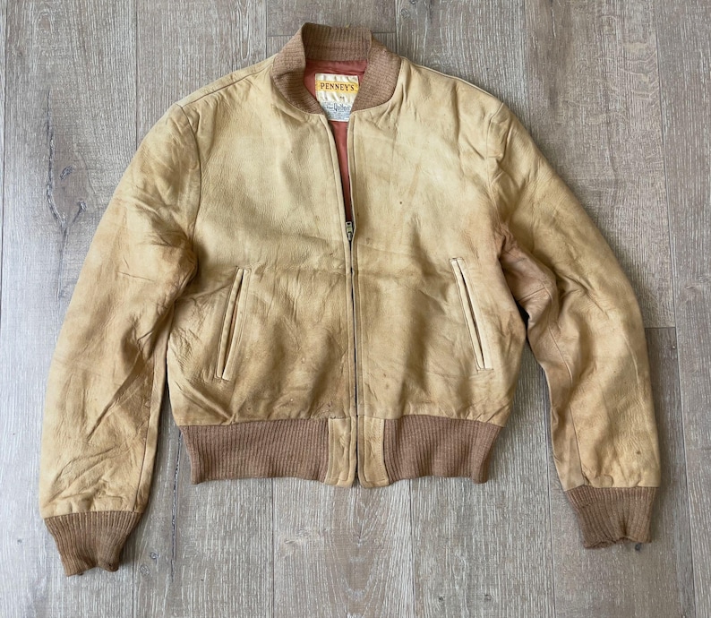 May include: A vintage tan leather bomber jacket with a zip-up front, two front pockets, and brown ribbed cuffs and hem. The jacket has a faded appearance and a small tag that reads "PENNEY'S".