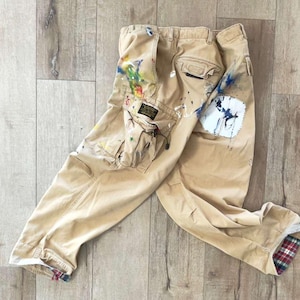 May include: Khaki cargo pants with paint splatters in various colors. The pants have multiple pockets and a patch on the knee. The cuffs are lined with a plaid pattern. The pants are laid out on a wooden surface.