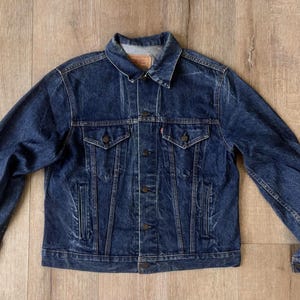 May include: A dark blue denim jacket with a button-up front, two chest pockets, and two side pockets. The jacket has a classic collar and long sleeves. A small red tag is visible on the chest pocket.
