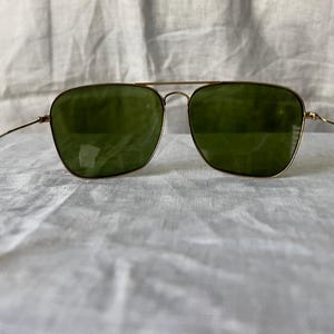 May include: Gold-framed sunglasses with green lenses. The glasses have a double bridge and thin temples. The lenses are square-shaped, and the frame is a warm gold color. The sunglasses are set against a white, textured background.