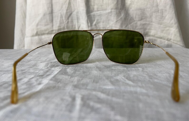 May include: Gold-framed sunglasses with green lenses. The glasses have a double bridge and thin temples. The lenses are square-shaped, and the frame is a warm gold color. The sunglasses are set against a white, textured background.