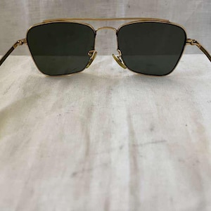 Puede incluir: Gafas de sol con montura dorada y lentes verde oscuro. Las gafas tienen un doble puente y patillas de color claro. Las lentes tienen una forma de aviador cl&aacute;sica. Sobre un fondo blanco.