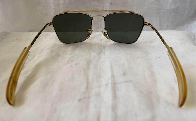 Puede incluir: Gafas de sol con montura dorada y lentes verde oscuro. Las gafas tienen un doble puente y patillas de color claro. Las lentes tienen una forma de aviador cl&aacute;sica. Sobre un fondo blanco.