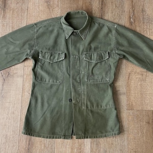 May include: A green utility shirt with a button-up front and two large pockets on the chest. The shirt has a pointed collar and long sleeves.