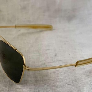 Puede incluir: Gafas de sol con montura dorada y lentes oscuras. Las gafas de sol de estilo aviador tienen un doble puente y puntas de patillas transparentes. Las gafas est&aacute;n en &aacute;ngulo, mostrando la forma de las lentes y la montura dorada.
