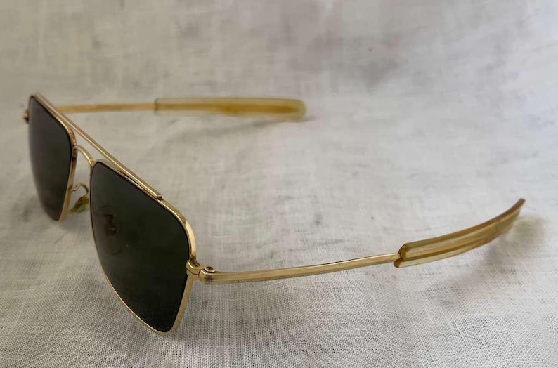 Puede incluir: Gafas de sol con montura dorada y lentes oscuras. Las gafas de sol de estilo aviador tienen un doble puente y puntas de patillas transparentes. Las gafas est&aacute;n en &aacute;ngulo, mostrando la forma de las lentes y la montura dorada.