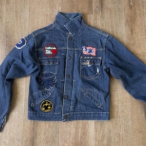 May include: A dark blue denim jacket with multiple patches. The jacket has a high collar, button closures, and two chest pockets. Patches include a California patch, a flag, and a scale of justice.