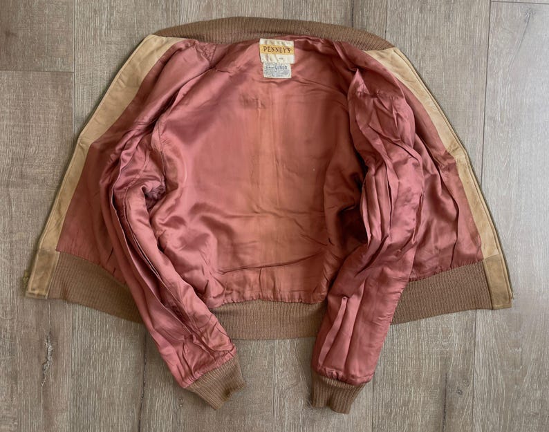 May include: A vintage brown suede jacket with a pink satin lining. The jacket features a ribbed collar and cuffs, and a tag that reads "PENNEYS". The jacket is laid flat, showcasing its interior and construction.
