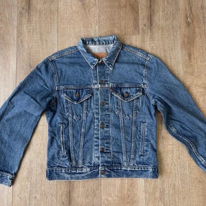 May include: A classic blue denim jacket with a button-up front, two chest pockets, and two side pockets. The jacket has a collared neckline and long sleeves. The denim is a medium wash with a slightly faded appearance.