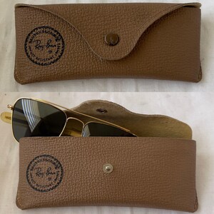 Puede incluir: Un estuche de gafas de cuero marr&oacute;n con cierre de bot&oacute;n a presi&oacute;n, con el logotipo de Ray-Ban. El estuche est&aacute; abierto, revelando gafas de sol con montura dorada y lentes oscuras.