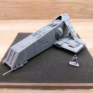 May include: A gray model of a spacecraft with detailed panels, wings, and a ramp. A small figurine stands nearby on a black surface. The model is displayed on a clear, rectangular base.