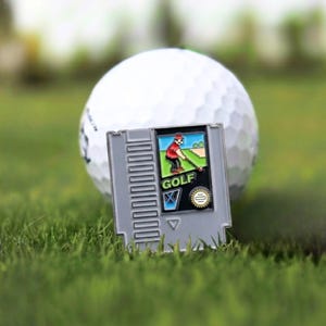 May include: A gray golf-themed video game cartridge pin with a colorful image of a golfer. The pin is set against a white golf ball and green grass, creating a playful contrast. The word "GOLF" is visible on the pin.