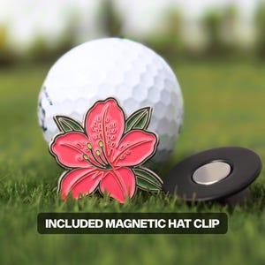 Könnte beinhalten: Ein Golfball liegt auf grünem Gras neben einem rot-grünen blütenförmigen Golfballmarker. Ein schwarzer magnetischer Hutclip ist ebenfalls enthalten. Der Text "INCLUDED MAGNETIC HAT CLIP" wird in einem schwarzen Rechteck angezeigt.