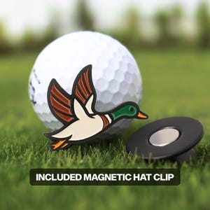 May include: A golf ball with a duck-shaped ball marker and a magnetic hat clip. The duck marker is white, brown, green, and orange. The hat clip is black with a silver magnetic center. The text "INCLUDED MAGNETIC HAT CLIP" is at the bottom.