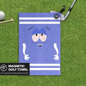 May include: A blue magnetic golf towel featuring a cartoon character with red eyes and a stressed expression. The towel has white stripes and the words "MAGNETIC GOLF TOWEL" are printed on a black rectangle. A golf club and ball are visible.
