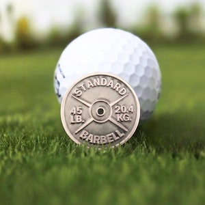 May include: A silver-colored barbell weight-shaped golf ball marker with the text "STANDARD BARBELL" and weight measurements of 45 LB and 20.4 KG. A white golf ball rests in the background on a green grassy surface.