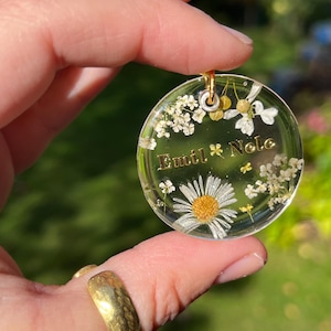 Può includere: Un ciondolo rotondo trasparente con fiori secchi e dettagli dorati. Il ciondolo presenta una margherita, piccoli fiori bianchi e dettagli in foglia d'oro. I nomi "Emil" e "Nela" sono scritti in lettere dorate.