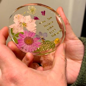 May include: A clear, round resin coaster with embedded dried flowers in shades of pink, purple, and yellow. Gold text includes names. A unique, handcrafted item for home decor.