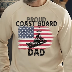 May include: Sand-colored sweatshirt with the text "Proud Coast Guard Dad" and a graphic of a ship over an American flag design. The sweatshirt is a crew neck style.