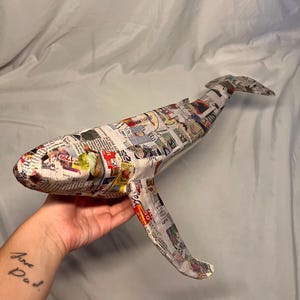 May include: A handcrafted whale sculpture made from recycled newspaper. The whale is covered in newsprint with visible text and images, and has fins and a tail. The sculpture is held in a hand against a light blue background.