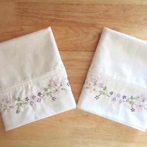 Op de afbeelding: Twee crèmekleurige kussenslopen met bloemenborduurwerk. Het borduurwerk toont kleine paarse en roze bloemen met groene stelen en bladeren. De kussenslopen zijn opgevouwen en op een houten oppervlak geplaatst.