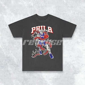 May include: Black t-shirt featuring a graphic of a basketball player in a red, white and blue jersey being wrapped by a snake with flames behind them. The text "PHILA" is above the graphic.