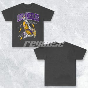 May include: Black t-shirt featuring a graphic print of a basketball player wearing a purple and gold jersey with the text "LOS ANGELES" above the player.