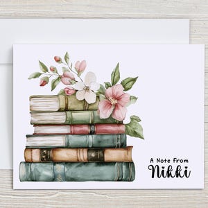 May include: A greeting card featuring a stack of books with watercolor floral accents. The card has a white envelope and the text "A Note From Nikki" in a handwritten font. The books are in shades of green, brown, and blue.