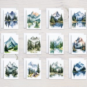 May include: A collection of watercolor greeting cards featuring mountain landscape scenes. Each card showcases a unique vista of mountains, trees, and water in shades of blue, green, and brown. The cards are displayed on a light wood surface.