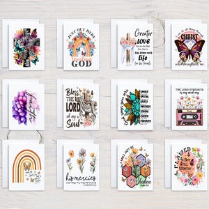 May include: A collection of white greeting cards with various designs. Some cards feature floral arrangements, butterflies, and religious text. Other cards have rainbow and cross designs. The cards are arranged on a light wood surface.