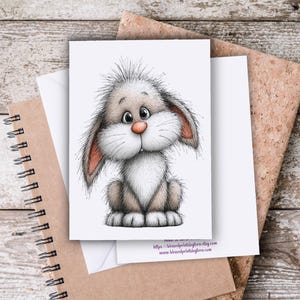 May include: A greeting card featuring a whimsical illustration of a fluffy bunny with large ears, a pink nose, and a soft, neutral color palette. The card is set against a wooden background with a spiral notebook and a corkboard.