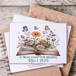 May include: A white greeting card with a floral design featuring an open book, flowers, and butterflies. The text "A New Chapter Begins Retired 2025" is printed in green. The card is on a wooden surface.