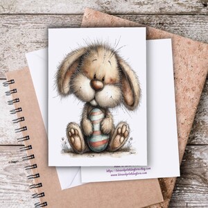 May include: A greeting card featuring a cartoon bunny holding a decorated Easter egg. The bunny has soft brown fur, large ears, and a round nose. The egg is striped with blue, red, and white. The card is on a wooden surface with a spiral-bound notebook and a corkboard.