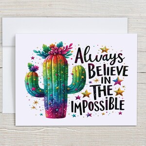 May include: Greeting card featuring a colorful cactus illustration with the text "Always Believe in the Impossible." The cactus is depicted in vibrant rainbow hues, adorned with stars and floral accents, set against a white background.