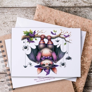 Puede incluir: Una tarjeta de felicitación blanca con un murciélago de dibujos animados colgado boca abajo de una rama, rodeado de murciélagos y arañas moradas. El murciélago tiene alas moradas, un sombrero de bruja y patas a rayas. La tarjeta está sobre una superficie de madera.