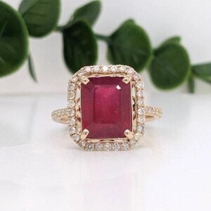 May include: A gold ring featuring a large, rectangular, deep red gemstone at its center. The stone is surrounded by a halo of small, clear, round-cut stones. The band is thin and metallic, with a polished finish that reflects light.
