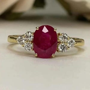 Puede incluir: Un anillo de oro con un gran rubí de corte ovalado como piedra central, flanqueado por grupos de piedras preciosas redondas y claras más pequeñas. El rubí es de color rojo intenso y el anillo tiene un diseño clásico y elegante. El anillo está engastado en una banda de oro.