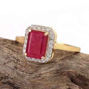 Può includere: Un anello in oro con una pietra centrale di rubino rosso a taglio smeraldo rettangolare. Il rubino è circondato da un alone di piccoli diamanti bianchi rotondi e chiari incastonati in una montatura d'oro. La fascia è in oro giallo lucido.
