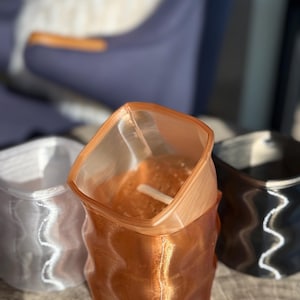 May include: Three 3D-printed, square-shaped vases in a row. The vases are in silver, copper, and black. The copper vase is in the center, with a wavy design. The vases have a translucent appearance, with a textured surface.