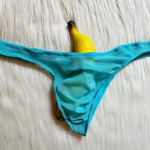 May include: A turquoise sheer thong with a banana inside, set against a white fluffy background. The thong has a low-rise design and a delicate, see-through fabric. The banana's yellow peel contrasts with the blue of the underwear.