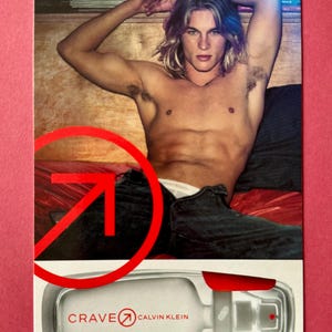 May include: A magazine advertisement featuring a shirtless person with long, light-coloured hair. The ad includes a bottle of "Crave" by Calvin Klein, with a red accent on the top. The background is a red surface.