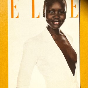 May include: Magazine cover featuring a woman in a white jacket. The word "ELLE" is printed in orange at the top. The woman has dark skin and a short haircut. The background is a light cream color.