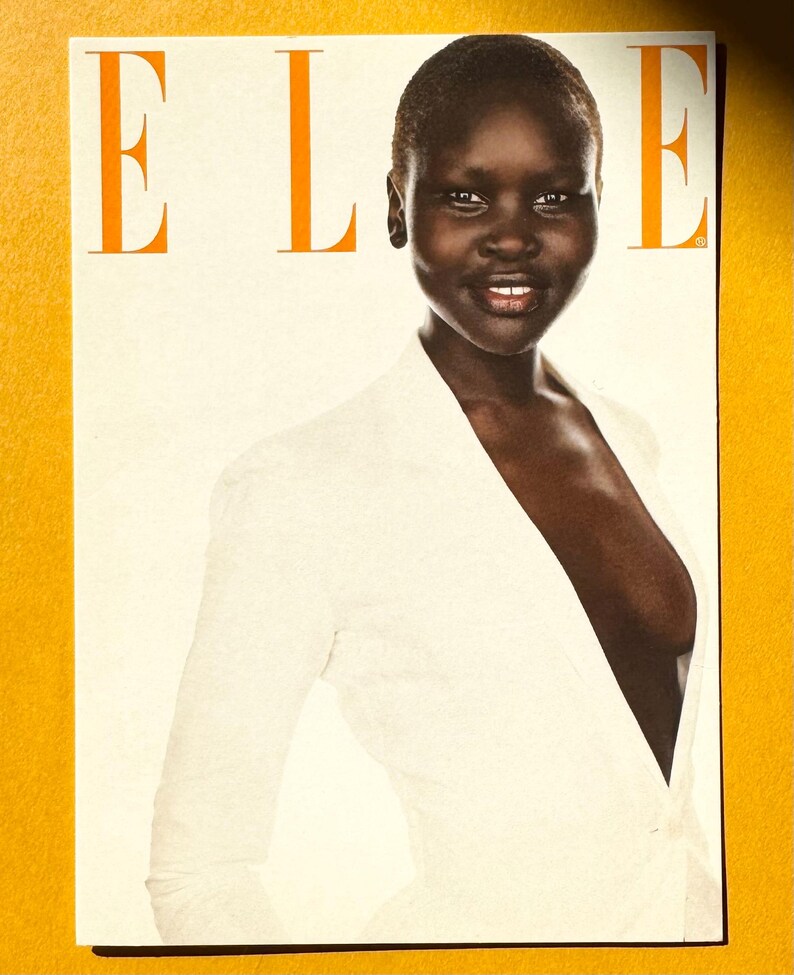 May include: Magazine cover featuring a woman in a white jacket. The word "ELLE" is printed in orange at the top. The woman has dark skin and a short haircut. The background is a light cream color.