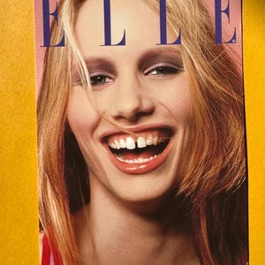 May include: A magazine cover featuring the word "ELLE" in large, blue capital letters. The cover image is a close-up of a woman with blonde hair, smiling widely. The background is a soft pink.