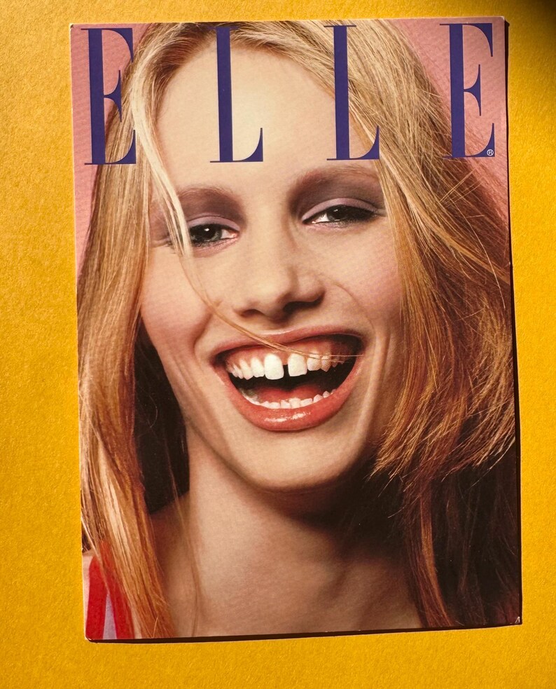 May include: A magazine cover featuring the word "ELLE" in large, blue capital letters. The cover image is a close-up of a woman with blonde hair, smiling widely. The background is a soft pink.