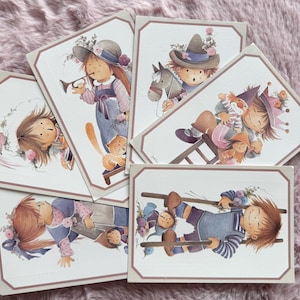 May include: A collection of vintage greeting cards featuring whimsical illustrations of children in various scenes. The cards showcase soft colors, detailed artwork, and a charming, nostalgic aesthetic. Each card has a decorative border.