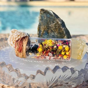 May include: A small glass bottle filled with dried flowers and herbs, a white selenite crystal, a grey labradorite crystal, and a clear quartz crystal on a clear glass plate with the words "UNKNOWN ON" inscribed on the plate.