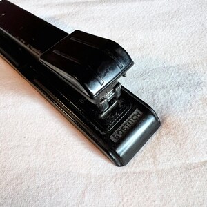 May include: A black Bostitch stapler, a common office supply, is shown in a close-up shot. The stapler is made of black plastic and metal. The word "BOSTITCH" is embossed on the base of the stapler.