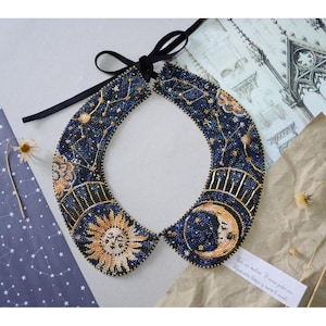 Puede incluir: Un collar de cuentas con diseño celestial que presenta el sol, la luna y las estrellas. El collar está hecho de cuentas azules, doradas y blancas y tiene un lazo de cinta negro.